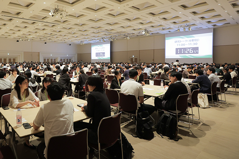 大ホールに長机が並び、多くの参加者がグループごとに机を囲んで熱心に話をする様子