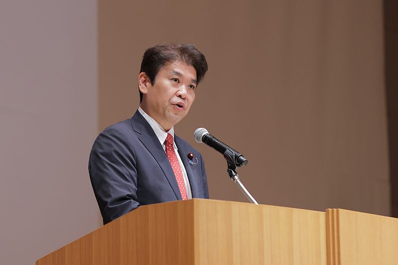 紺のスーツに赤いネクタイを締めた松本洋平文部科学大臣が、マイクの前で前を見据えて真剣に話す上半身のカット
