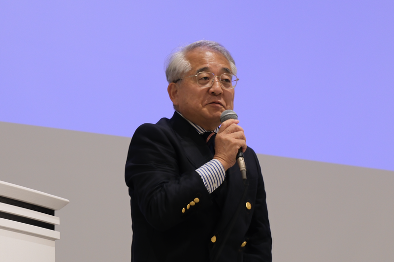 マイクを持って聴衆に向けて話す杉野理事長