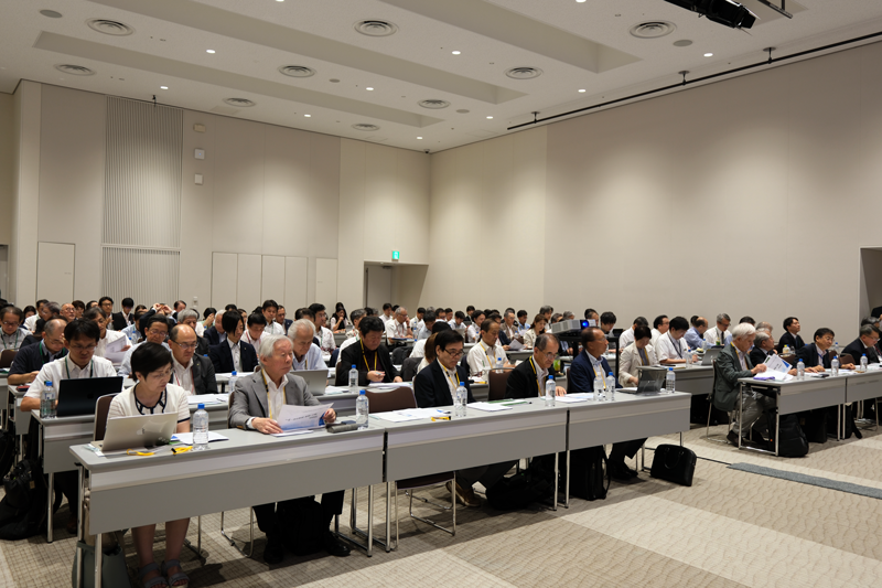 広い会議室の前方で行われている発表を聴く参加者たち